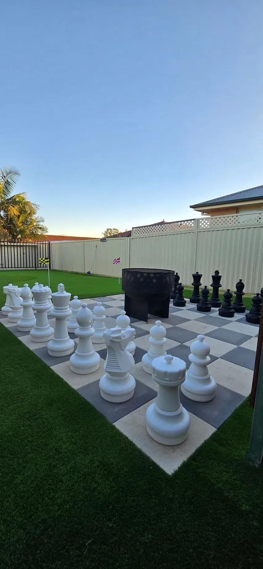 Giant chess set on a checkered patio, with white and black pieces. Green lawn and blue sky in the background.