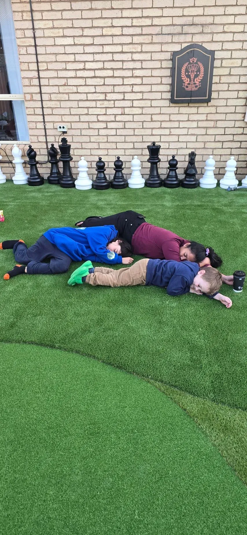 Three people lie on a green lawn, next to giant chess pieces, a brick wall is behind them.