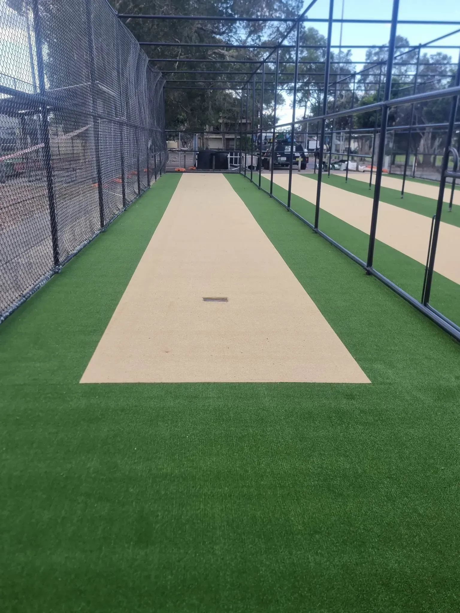 Cricket pitch with green artificial grass and beige playing surface, fenced in.
