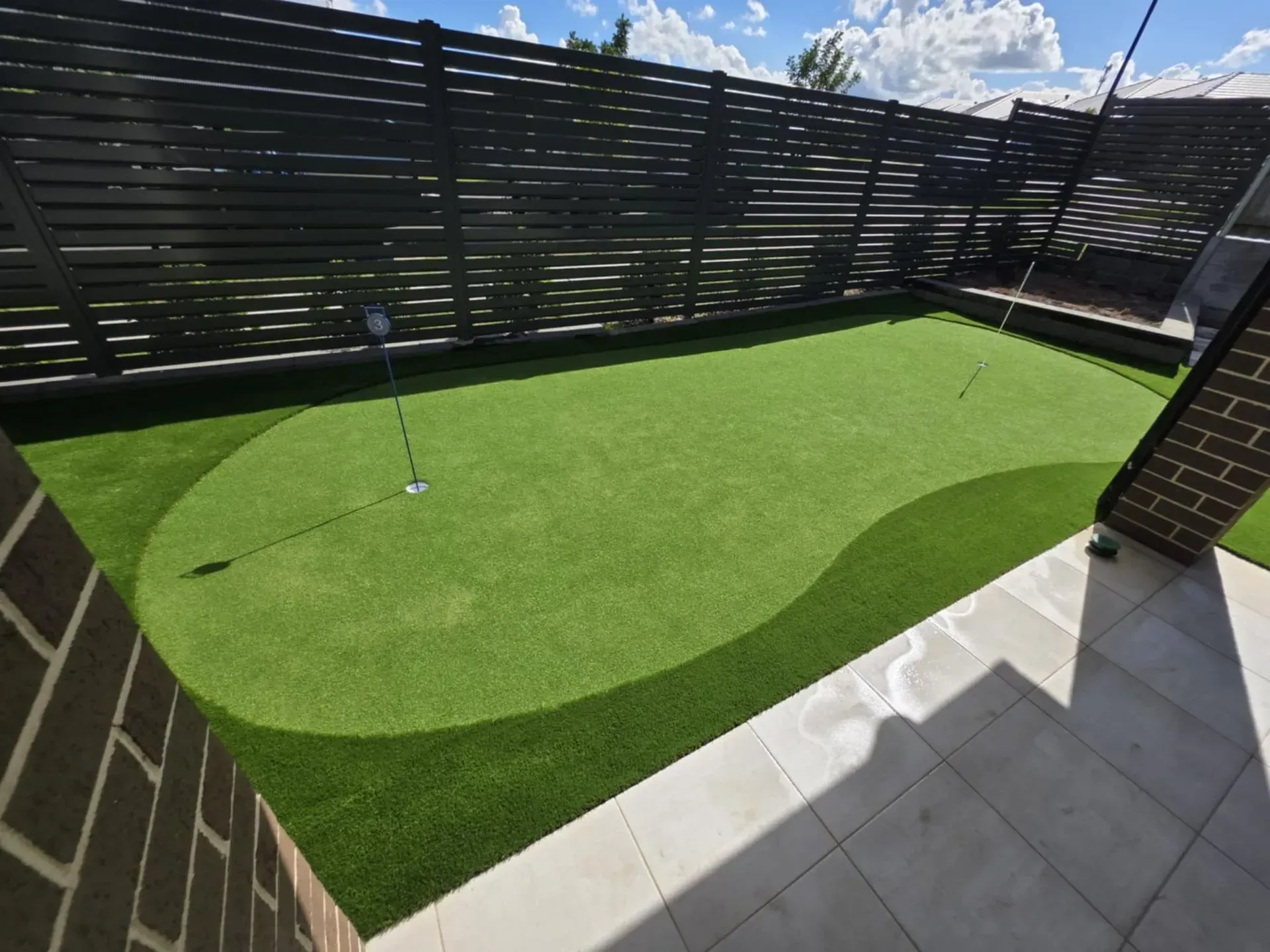 Backyard putting green with flags, surrounded by a dark fence and brick wall, under a blue sky.