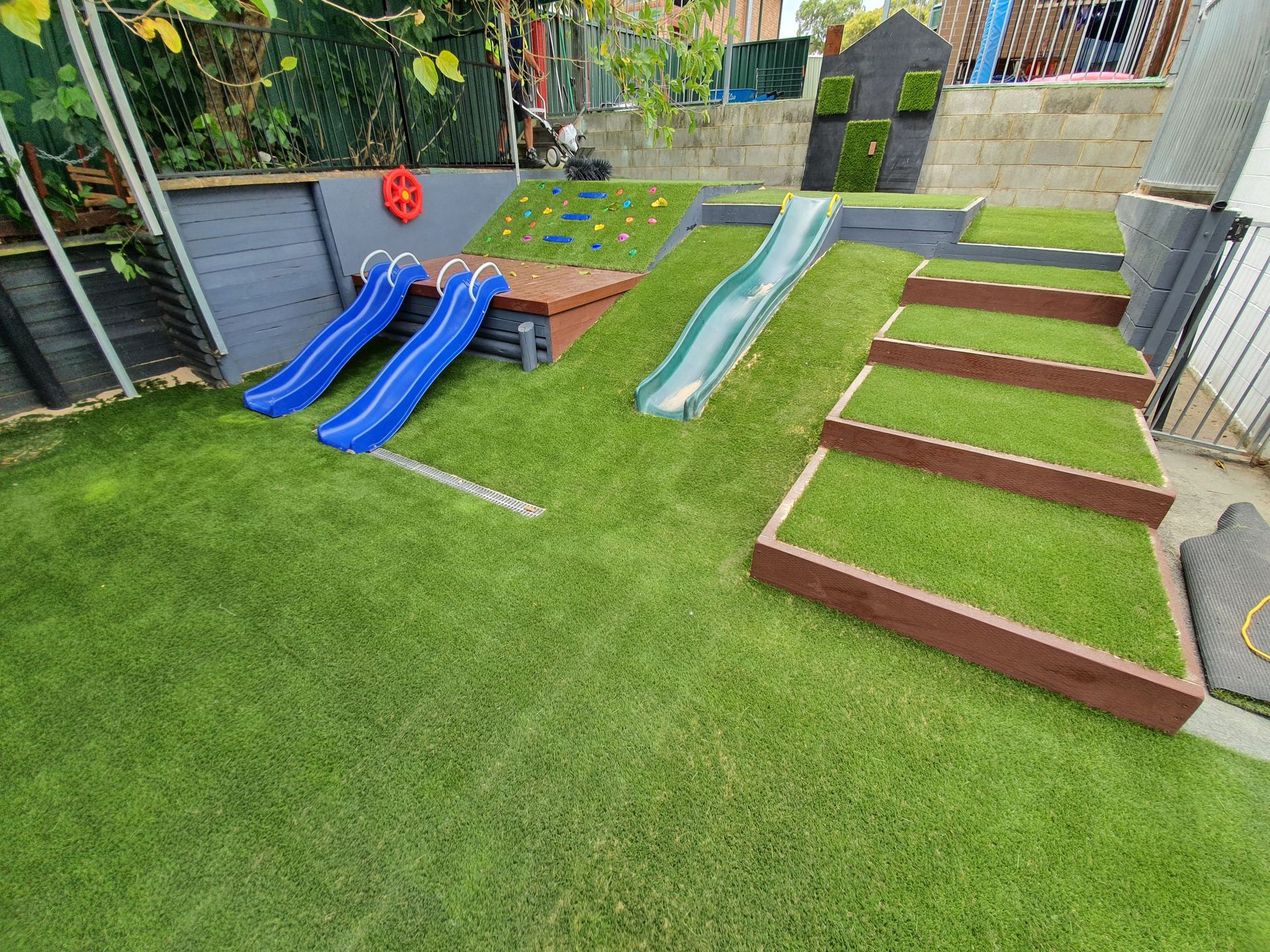 Playground with blue slides, green slide, climbing wall, and steps, all on artificial turf.