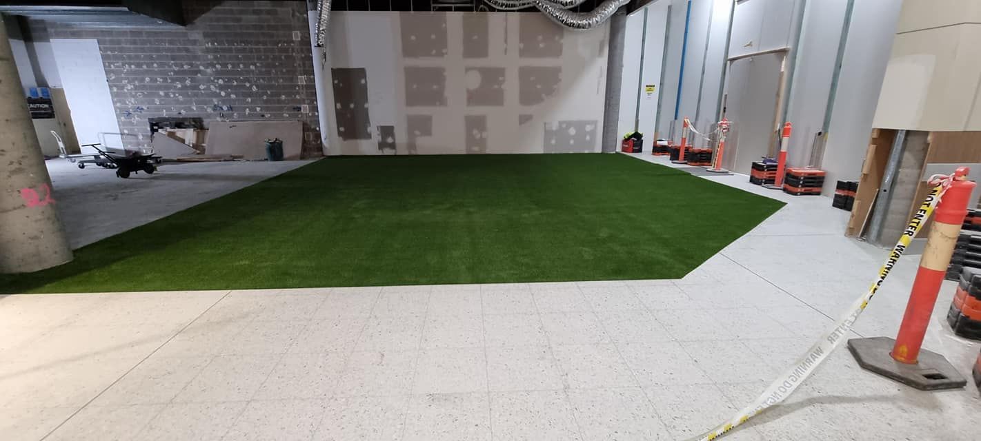 A partially installed artificial turf field in a construction zone. Orange traffic cones and building materials surround.