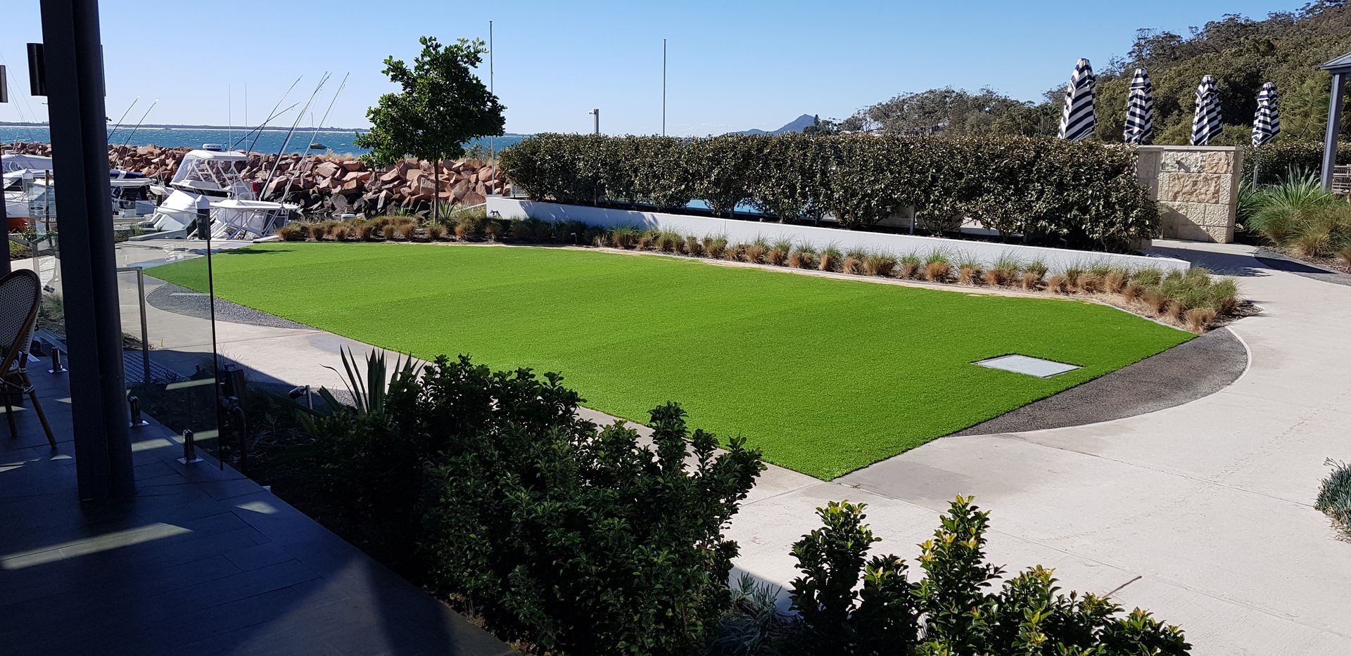 A green lawn near a waterfront with boats, bushes, and a pathway under a blue sky.