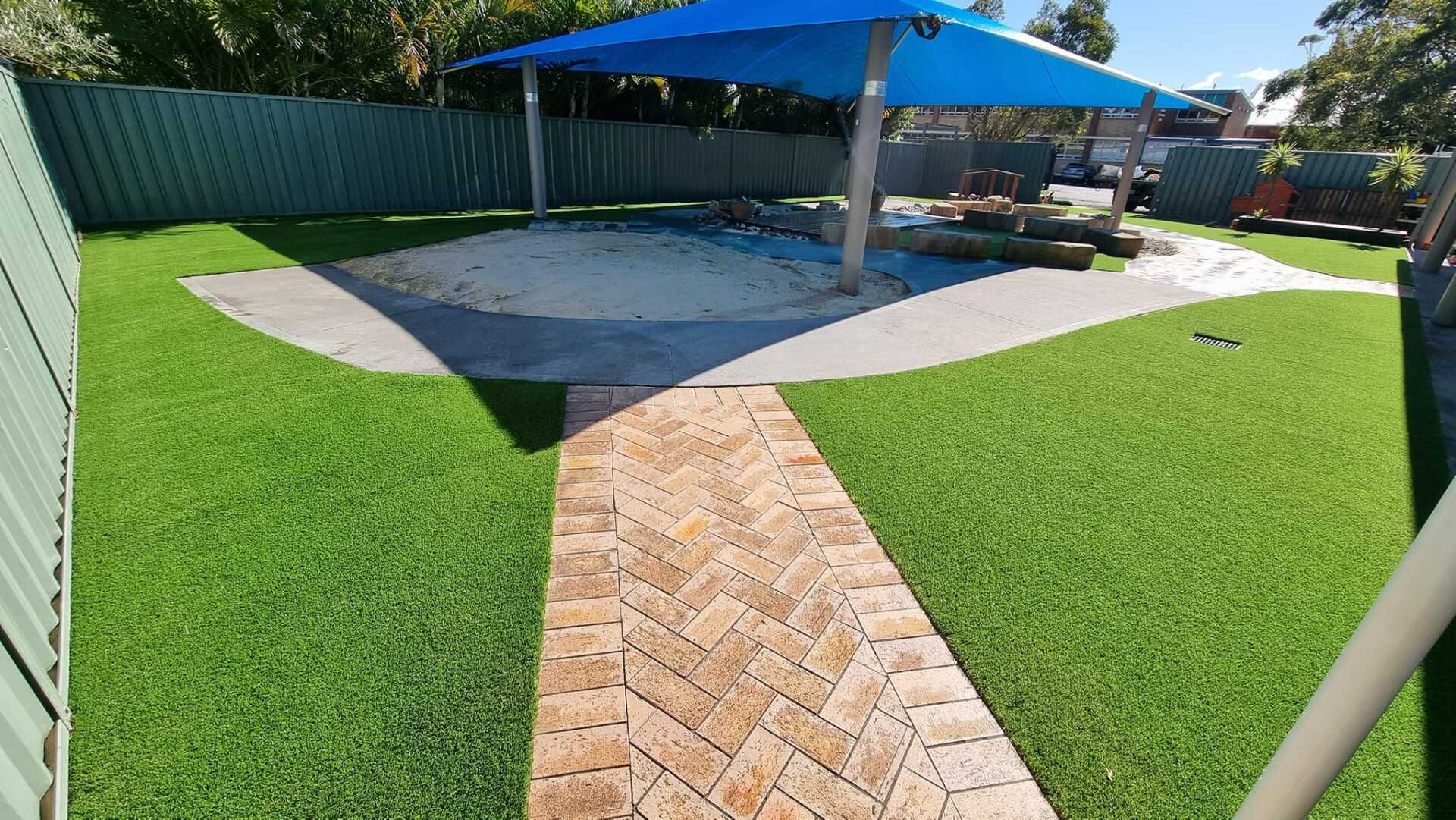 Playground with brick path, artificial grass, sand pit, and blue shade structure.