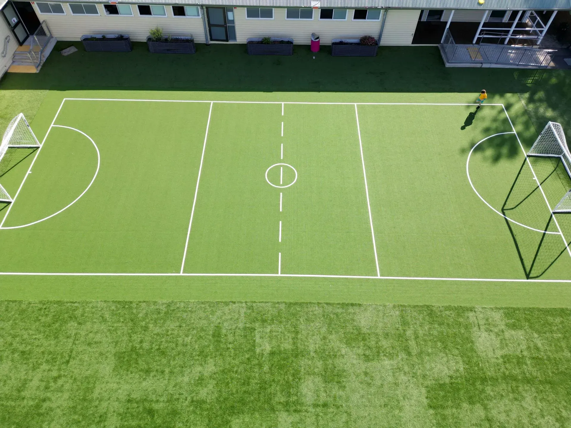 Overhead view of a small soccer field with white lines on green turf, goals at each end, and a building in the background.