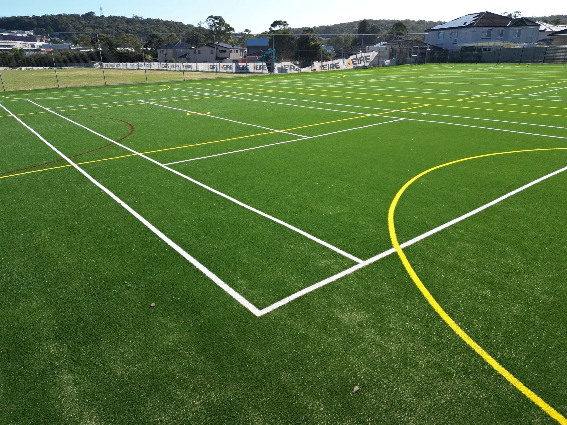 Green artificial turf field with white and yellow lines, buildings and trees in the background.
