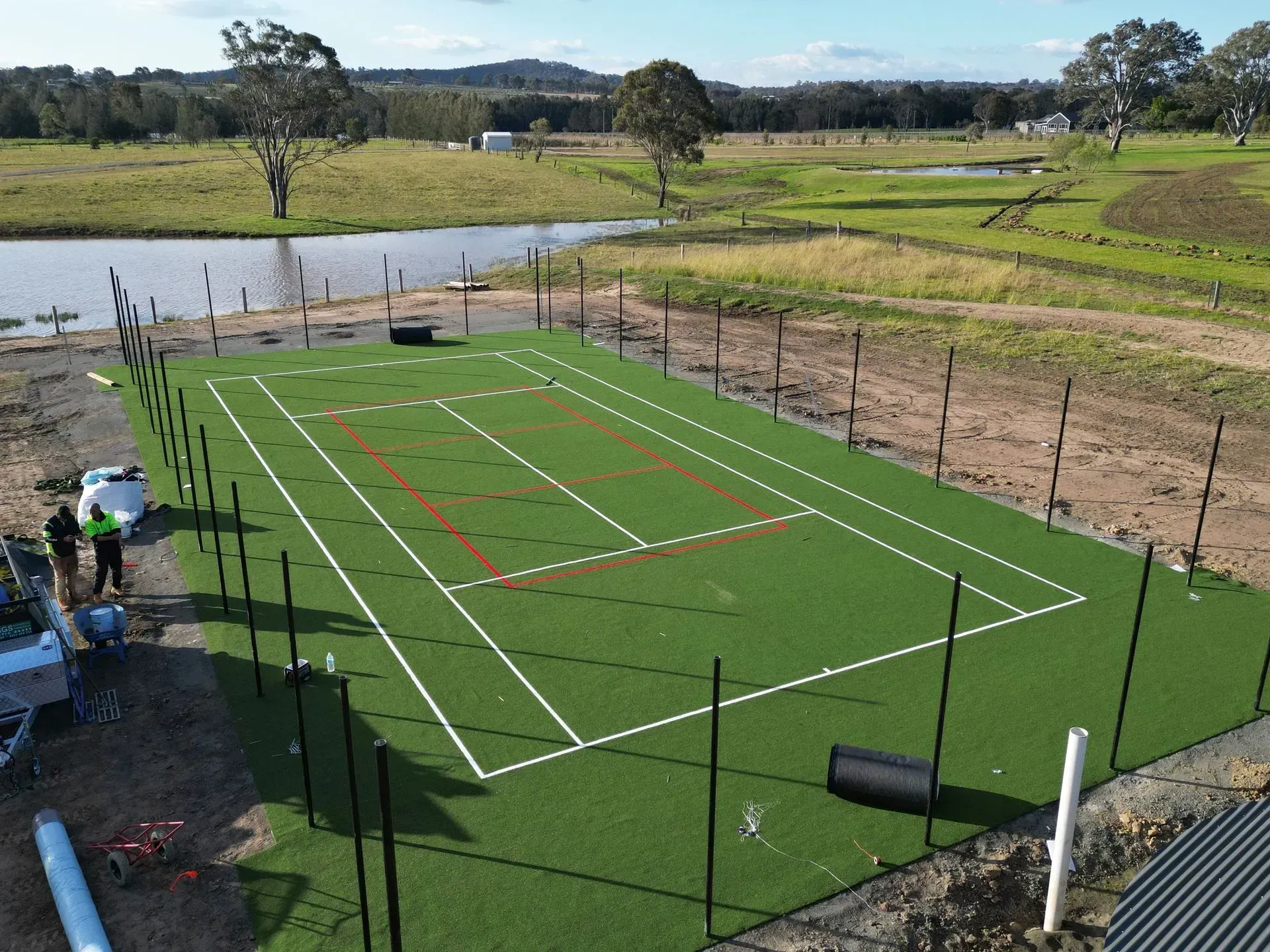 Aerial view of a green artificial turf tennis court with white and red lines, surrounded by black posts and a pond.