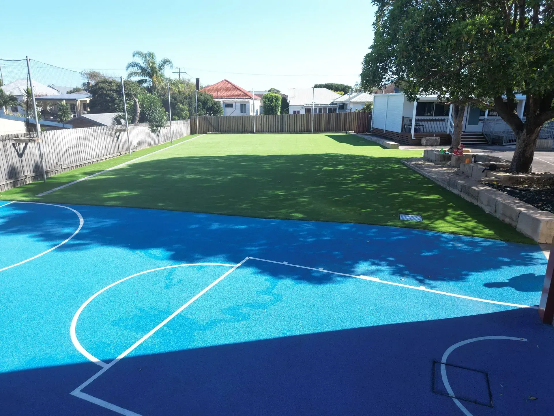 Blue basketball court and green turf playground in an outdoor setting with buildings in the background.