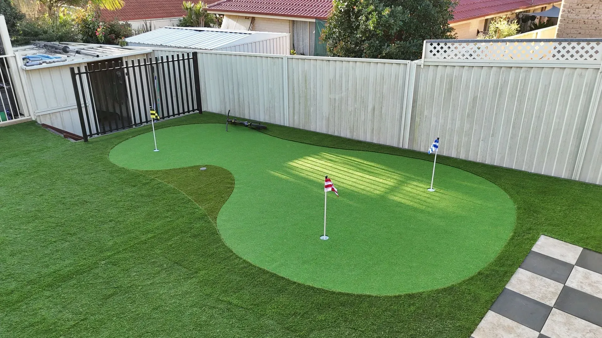 Backyard putting green with three flags, surrounded by artificial turf and a white fence.