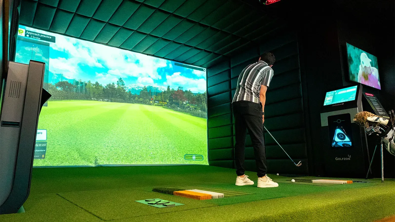 Person golfing in an indoor simulator. Green artificial turf, large screen showing course, black walls, monitors.