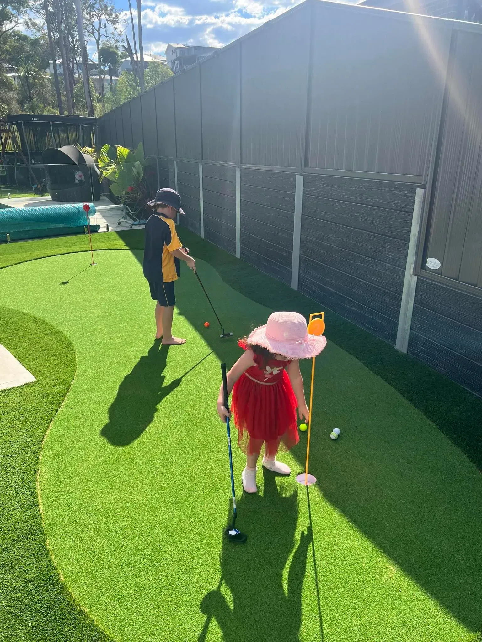 Two children play golf on a green lawn; one in a dress and hat, the other in a uniform, near a fence and pool.