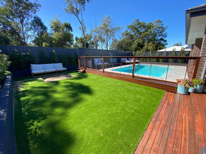 Backyard with artificial green grass, pool, wooden deck, sofa, and trees under a blue sky.