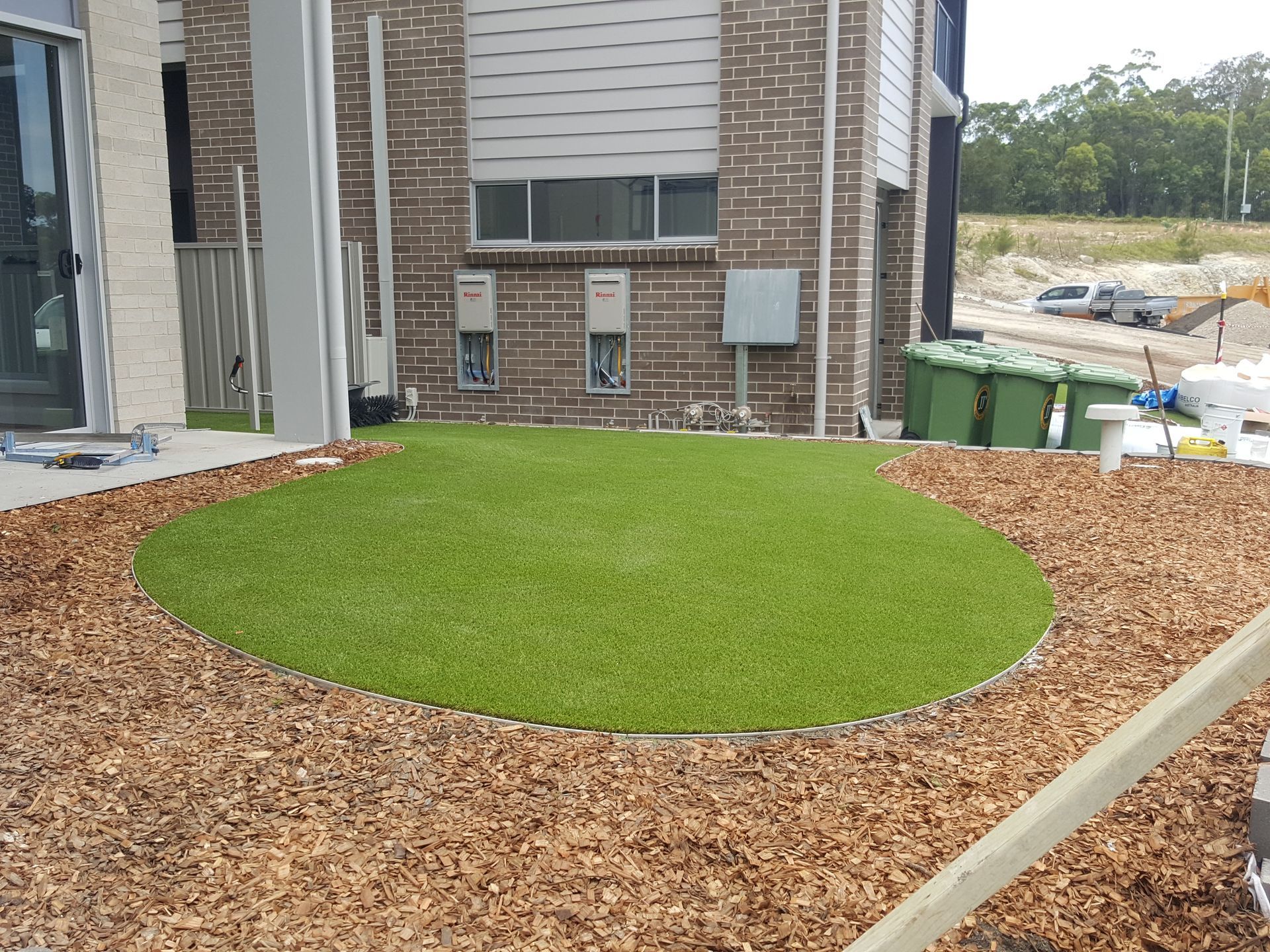A patch of artificial green turf surrounded by wood chips, near a brick building.