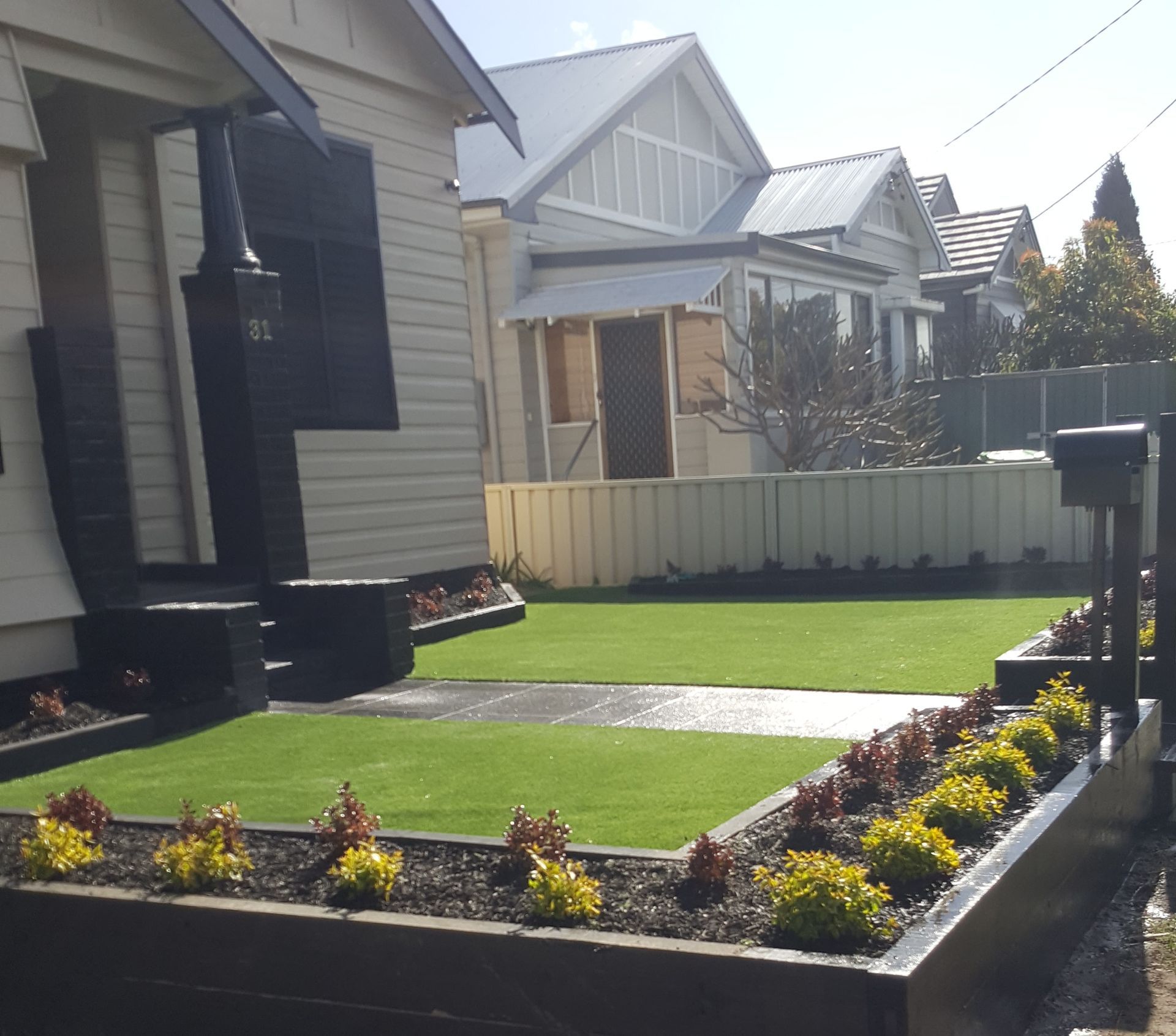 Front yard with artificial turf, raised garden beds with plants, and houses in the background.
