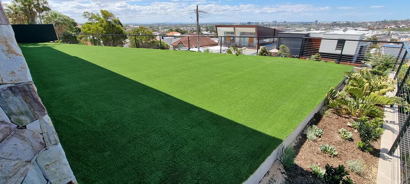Green artificial turf lawn with a cityscape view. There's a stone wall on the left and a garden bed on the right.