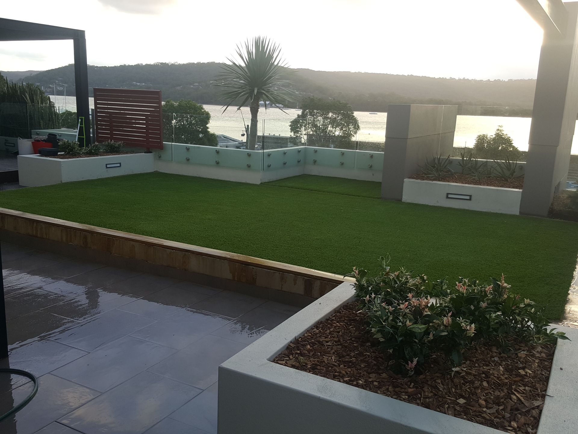 Rooftop patio with green lawn, planters, glass railings, and water view at dusk.