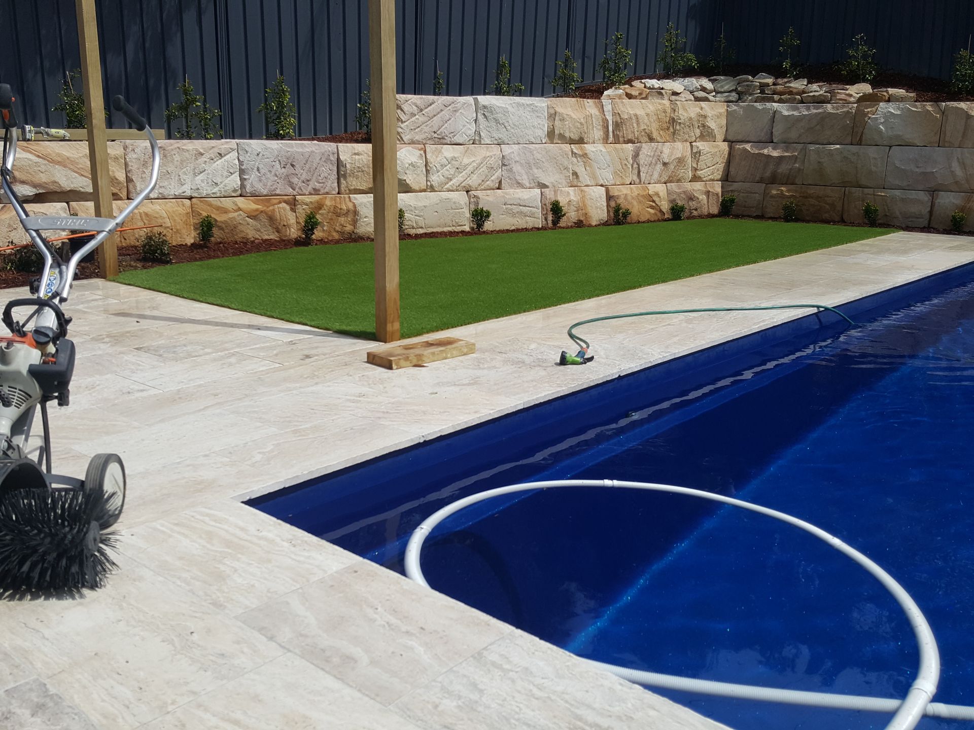 Poolside view with a small patch of artificial turf and a sandstone retaining wall. A pool cleaner and hose are present.