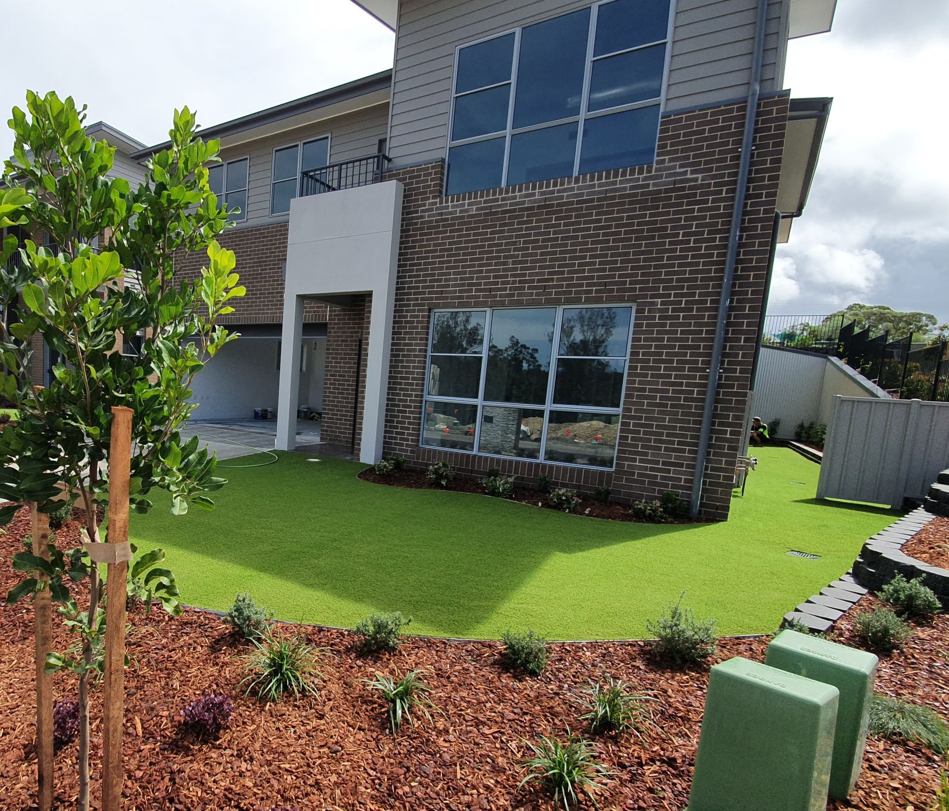 Two-story modern home with brick facade, large windows, and artificial green lawn, with landscaping in the front.