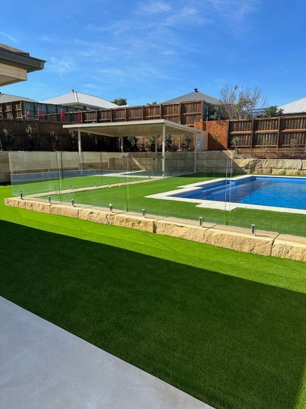 Green artificial turf surrounds a pool with glass fencing, a light brown retaining wall, and a shaded patio under a blue sky.