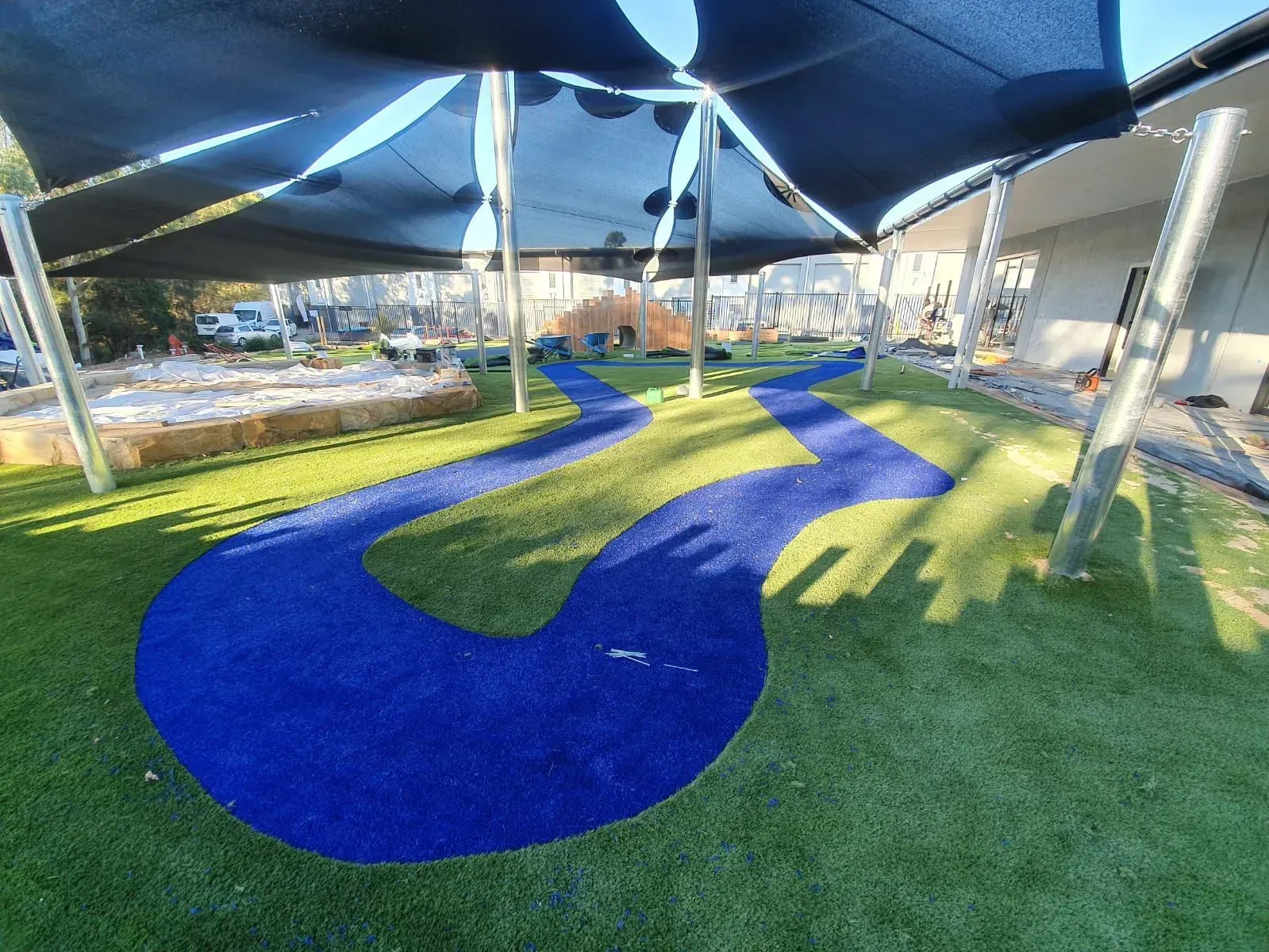 Playground with blue river-shaped path on green turf under a sunshade.