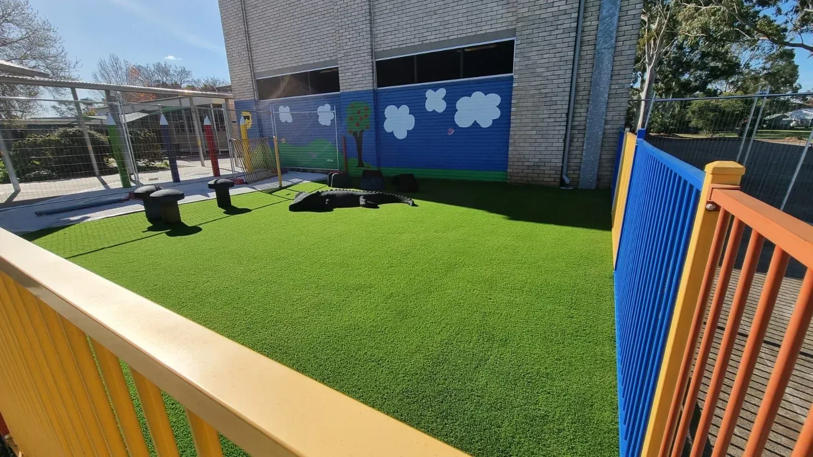 Playground with artificial turf, blue sky mural, and yellow and blue fencing.