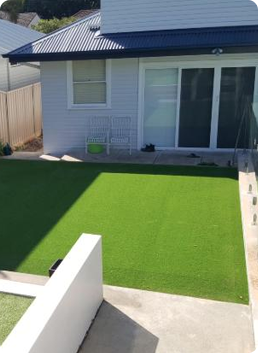 Backyard with green artificial turf, white house, sliding glass doors, concrete pathway.