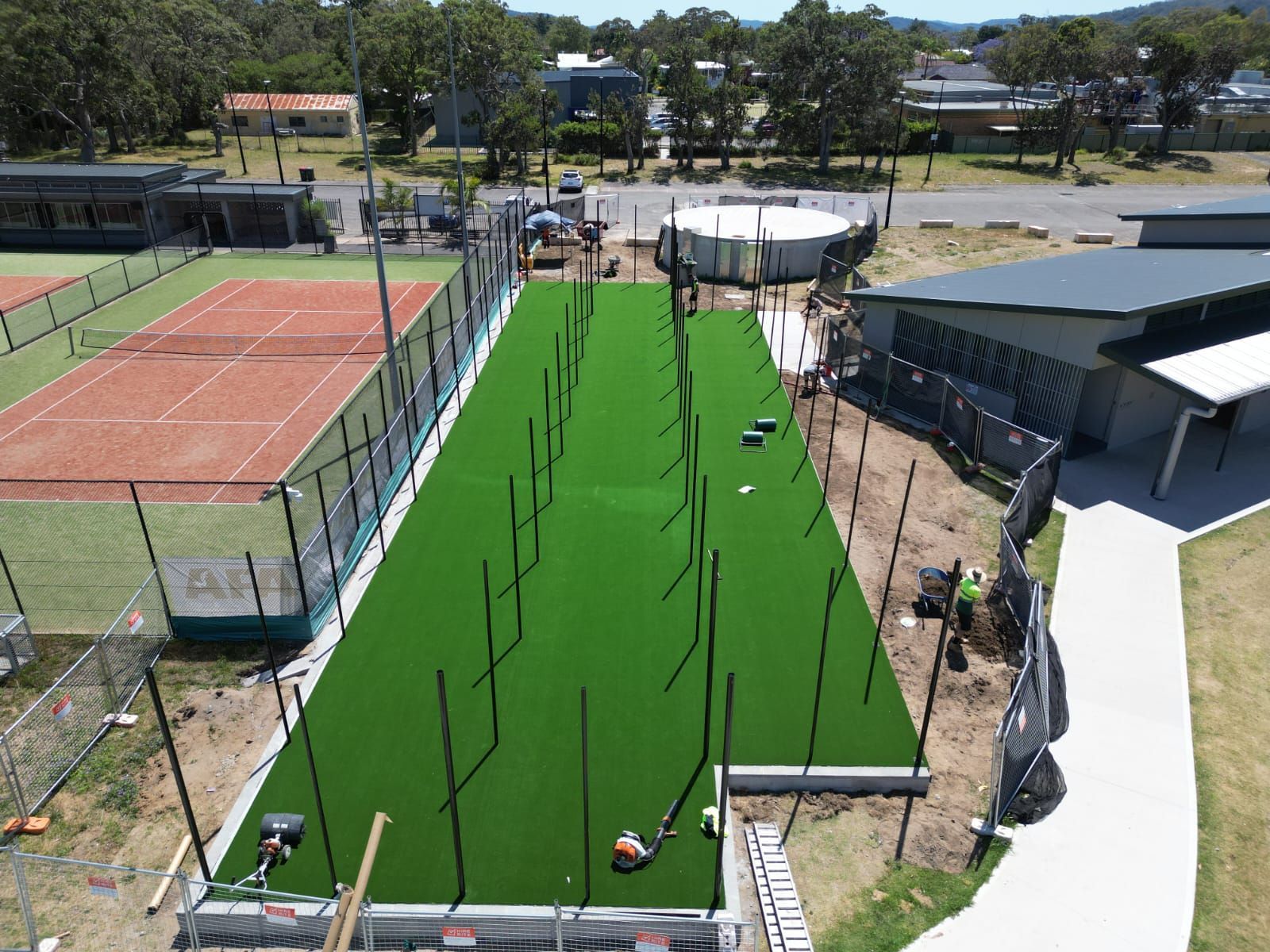 Aerial view of a sports field under construction with green turf, surrounding fence, and nearby buildings.