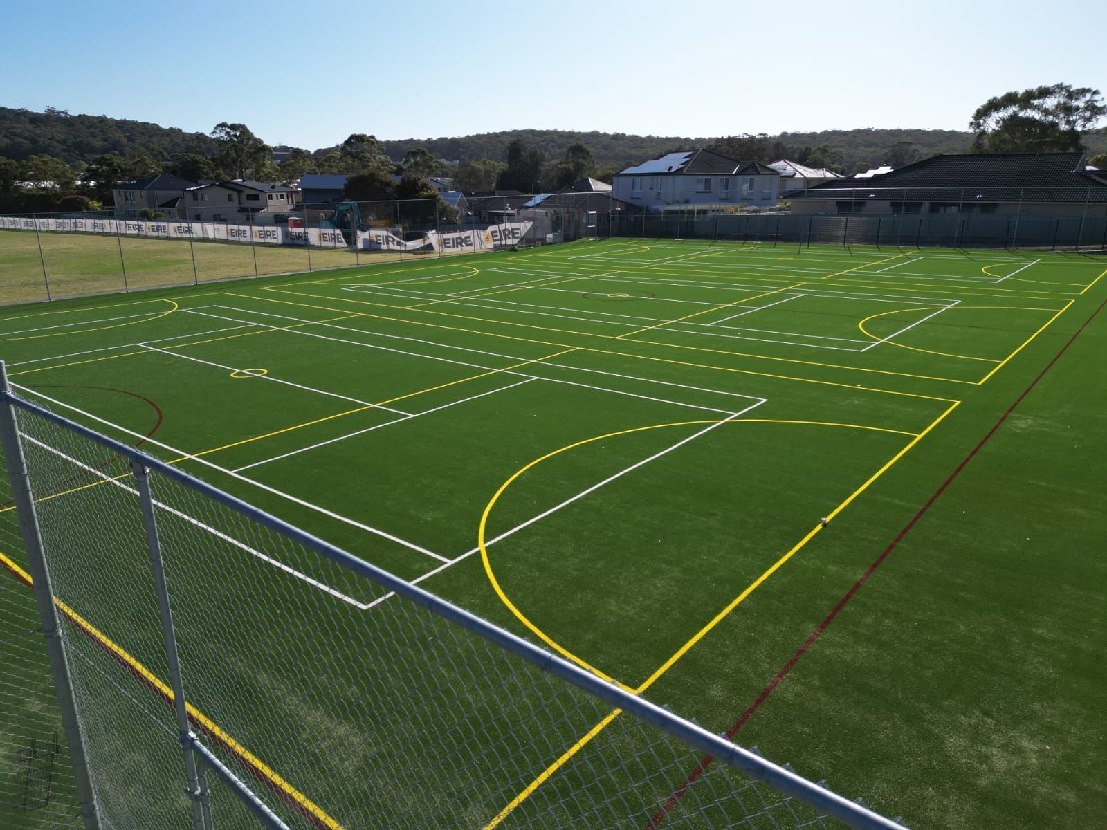 Green artificial turf sports field with white, yellow, and red markings, fenced perimeter, and buildings in the background.