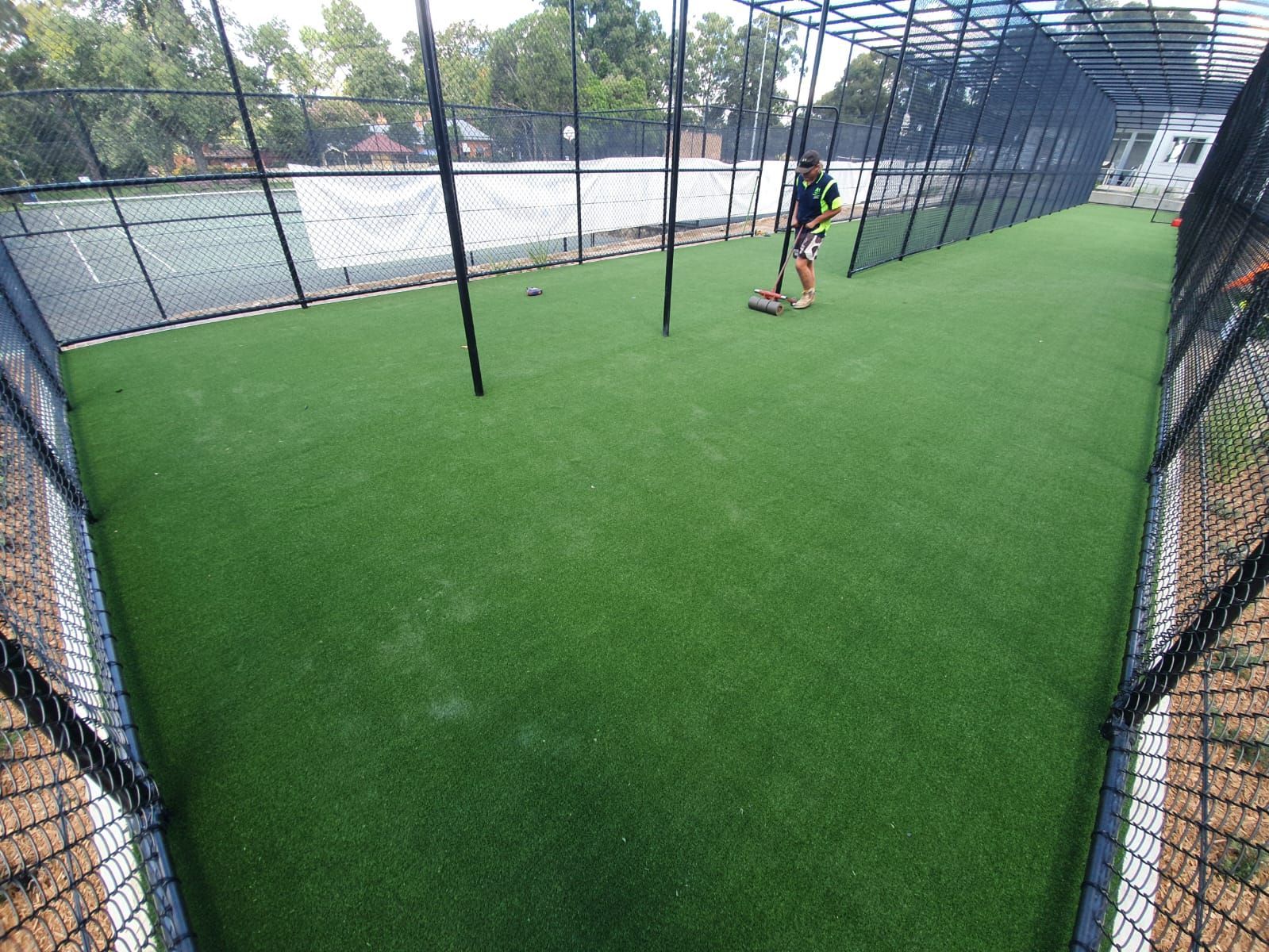 Green turf enclosed by netting; person stands inside, holding a scooter; tennis court visible in the distance.
