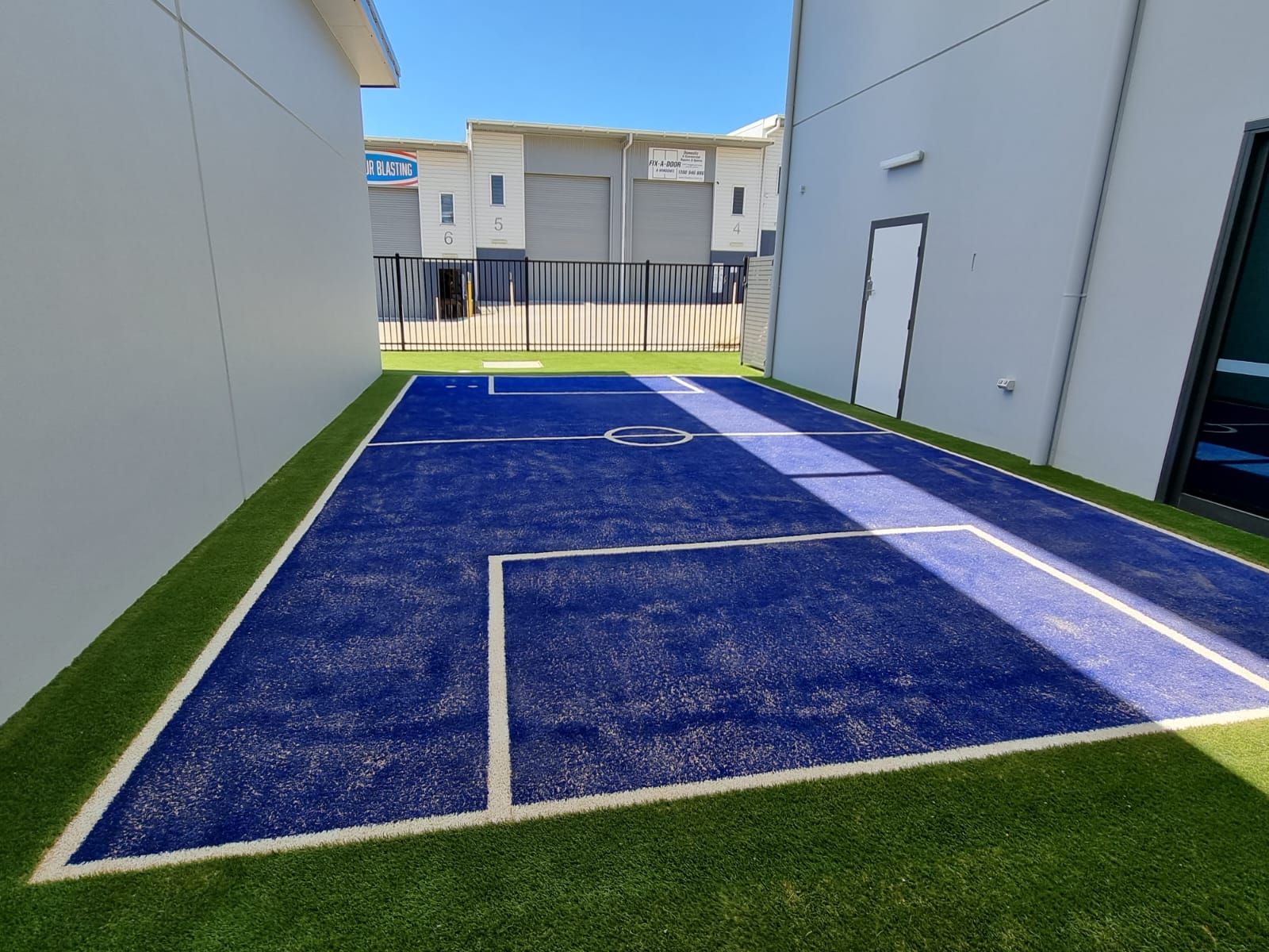 A blue court with white lines between two buildings; green artificial grass borders.
