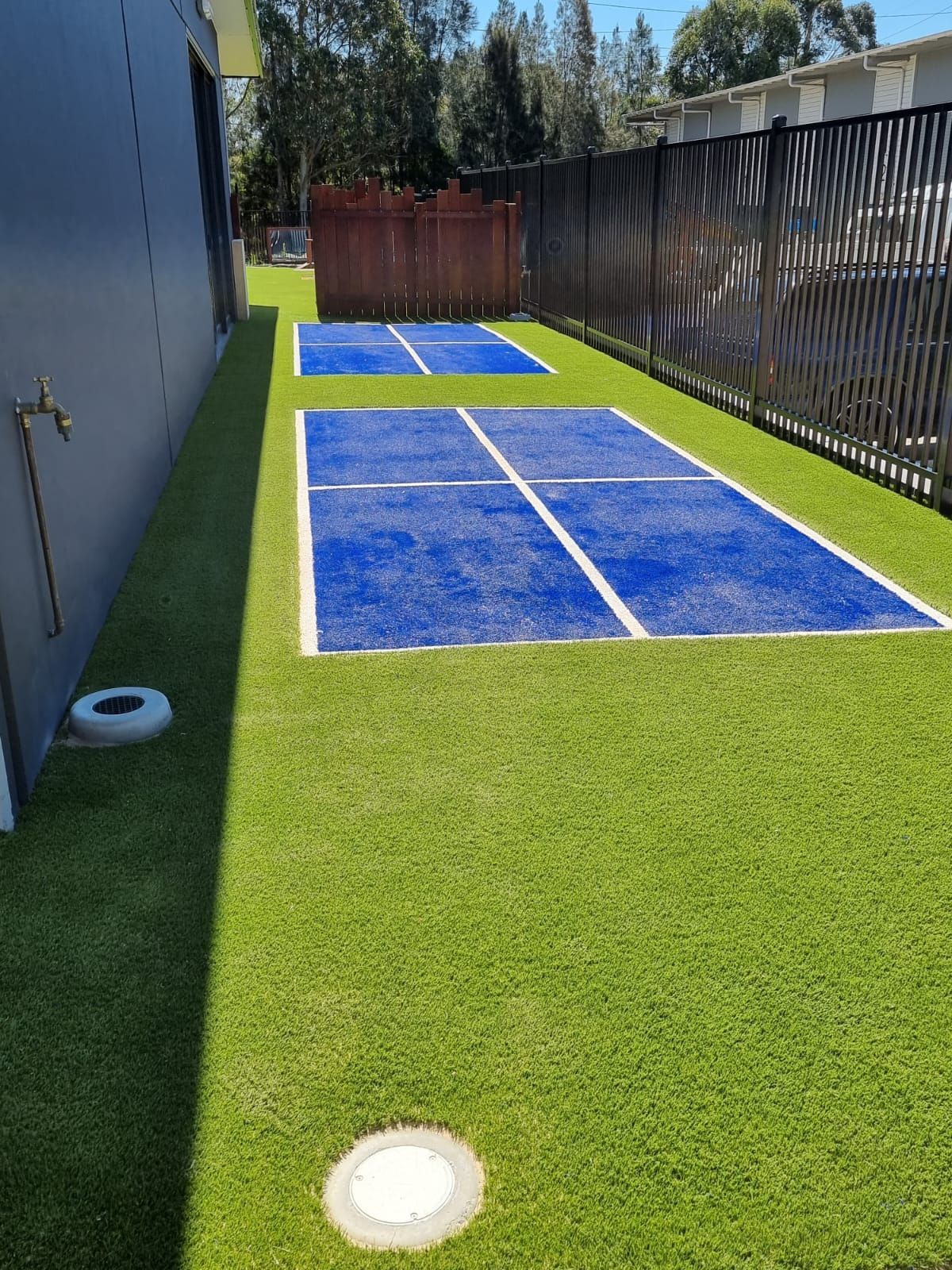 Green turf surrounds two blue shuffleboard courts with a brown fence and a dark building.