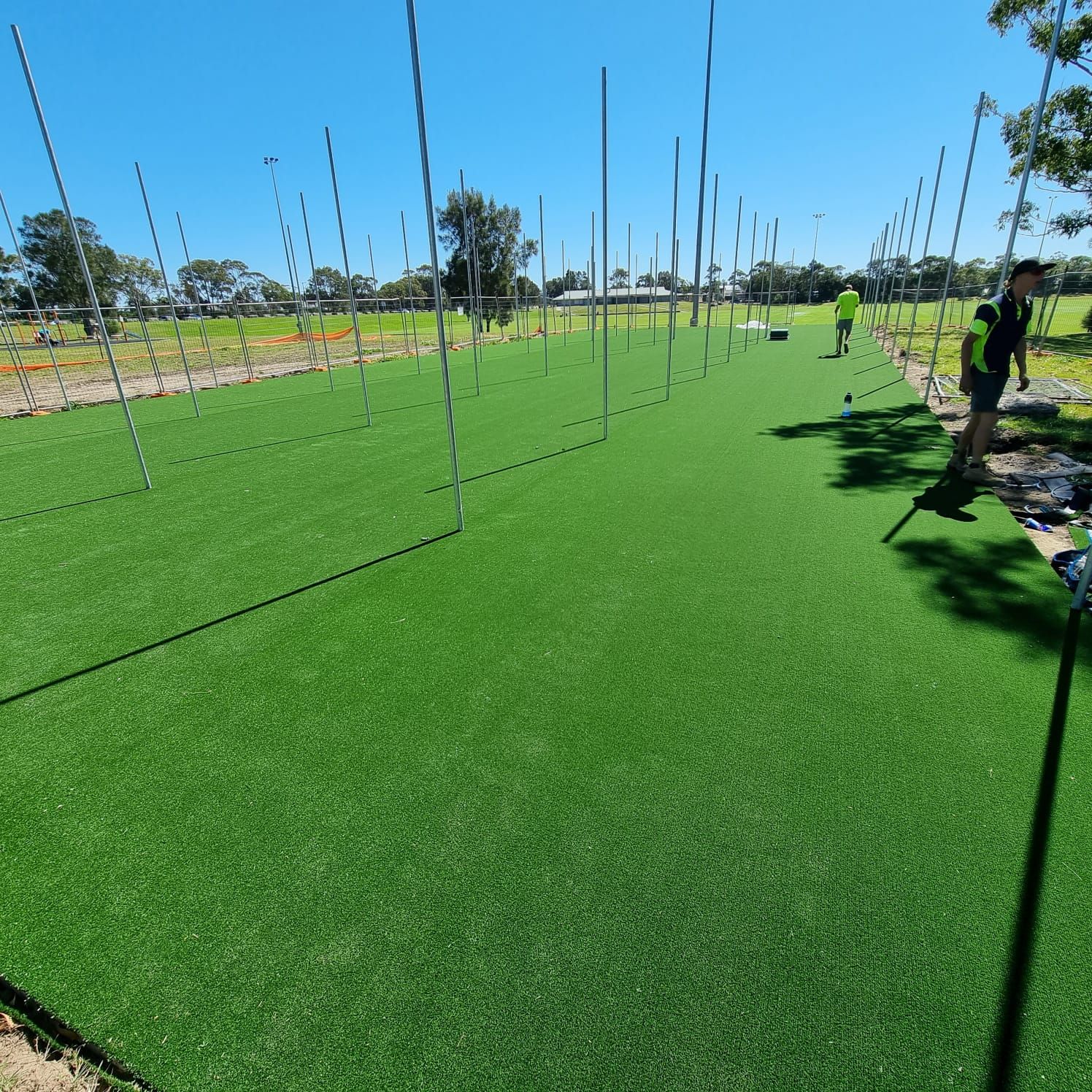 Artificial turf field being installed with workers and metal posts on a sunny day.