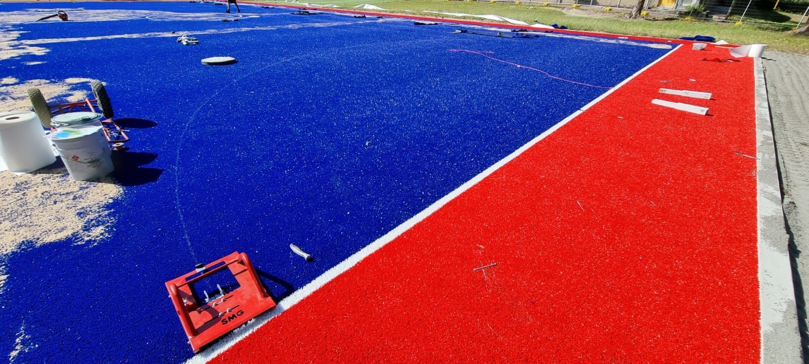 A track and field surface being installed, with red and blue sections. Buckets and tools are on the sandy area.
