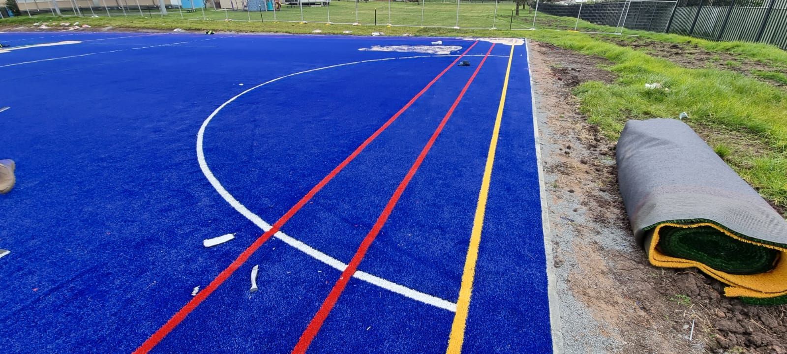 Blue sports field with white, red, and yellow markings, a roll of artificial turf is in the foreground.