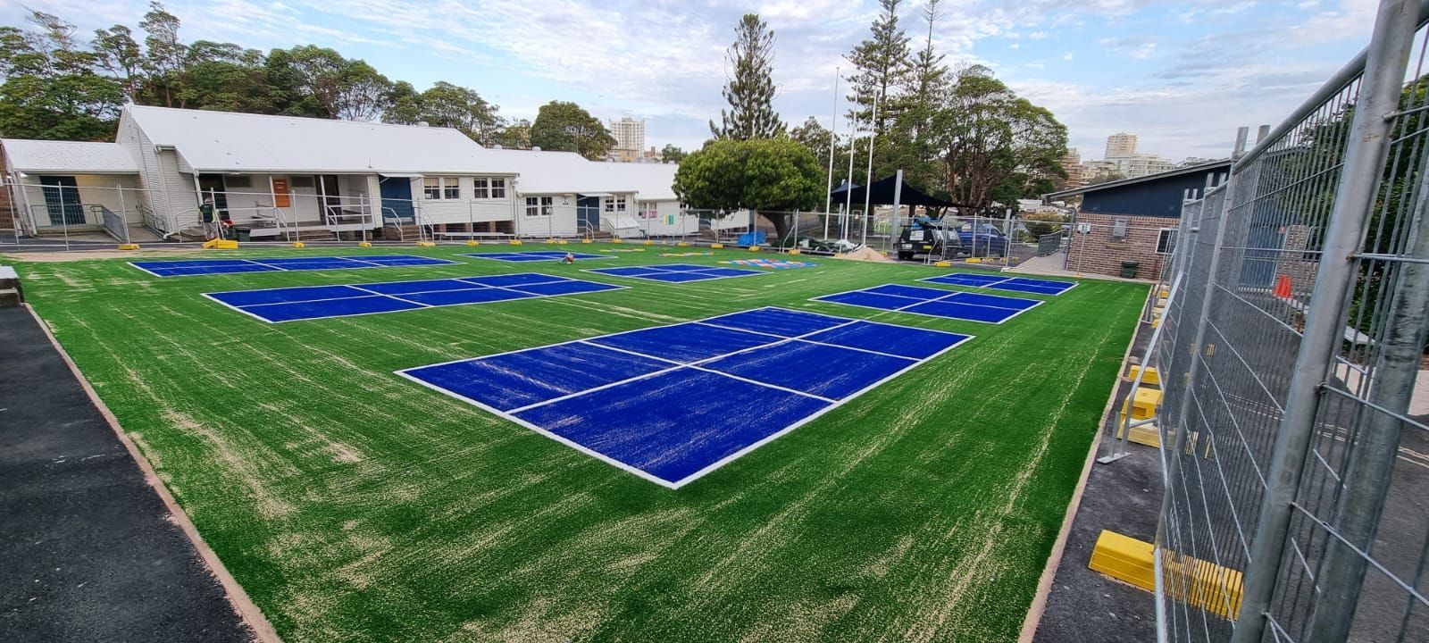 Newly installed green and blue artificial turf field with a building in the background. Construction fence in the foreground.
