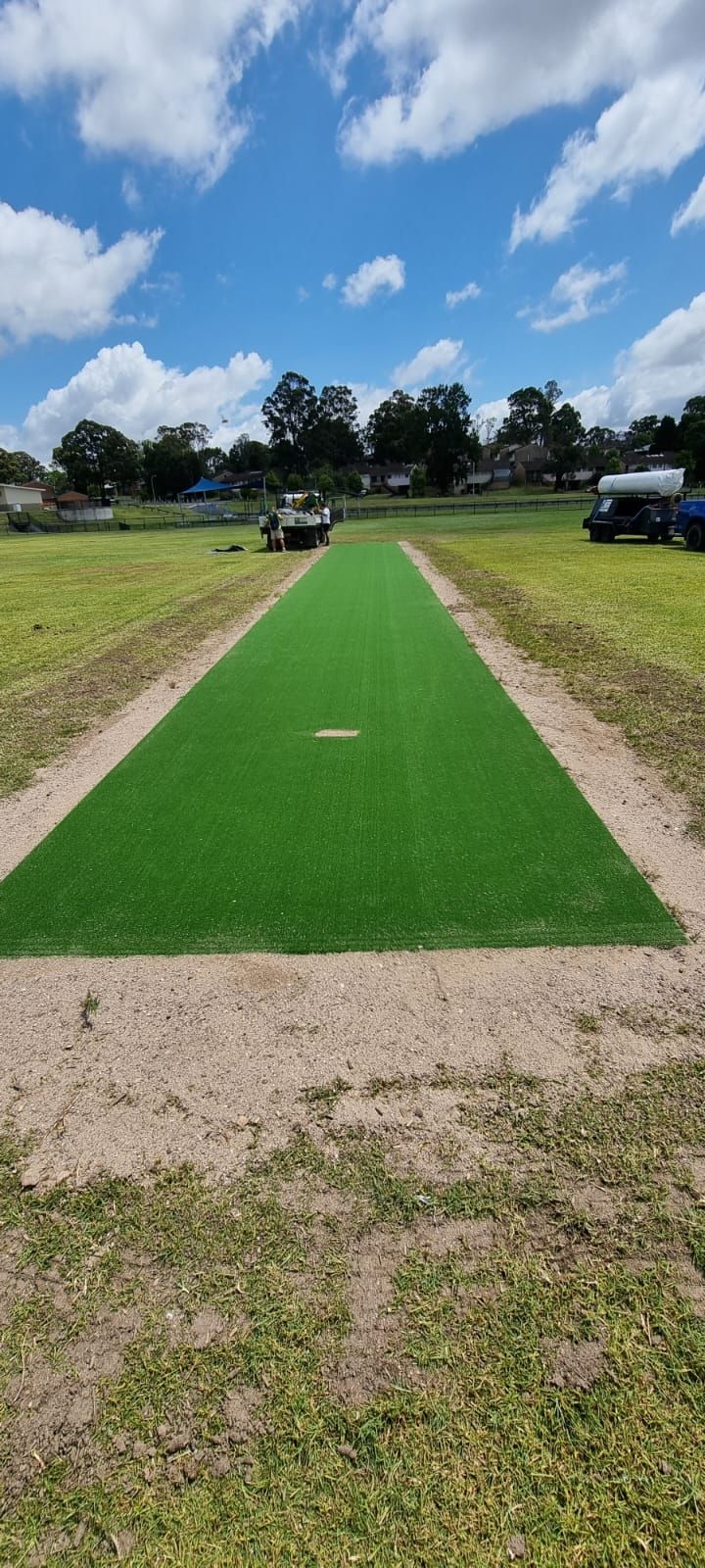 Green cricket pitch on a grassy field under a partly cloudy sky.