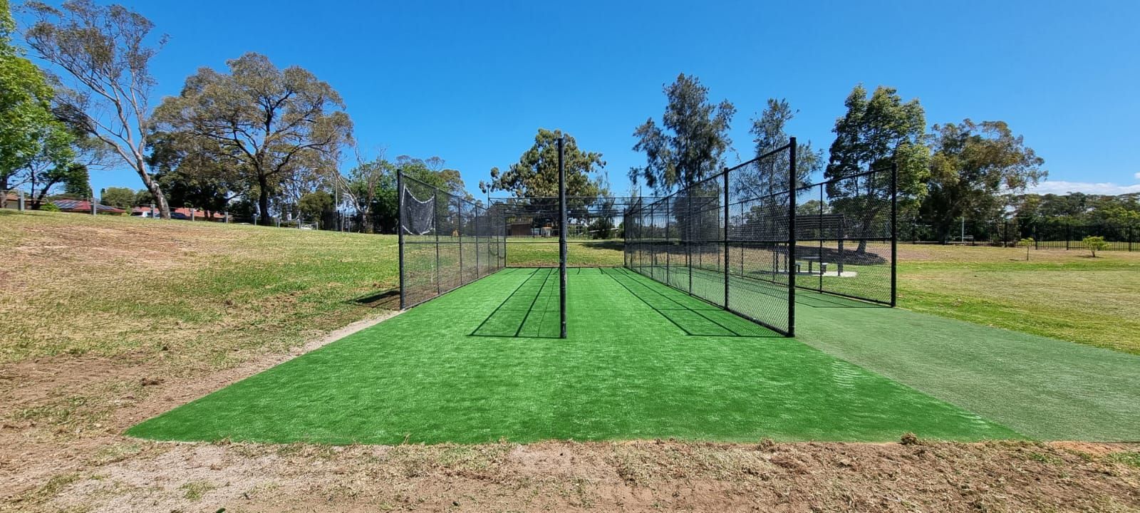 A cricket pitch in a park with artificial turf, surrounded by netting, trees and a blue sky.