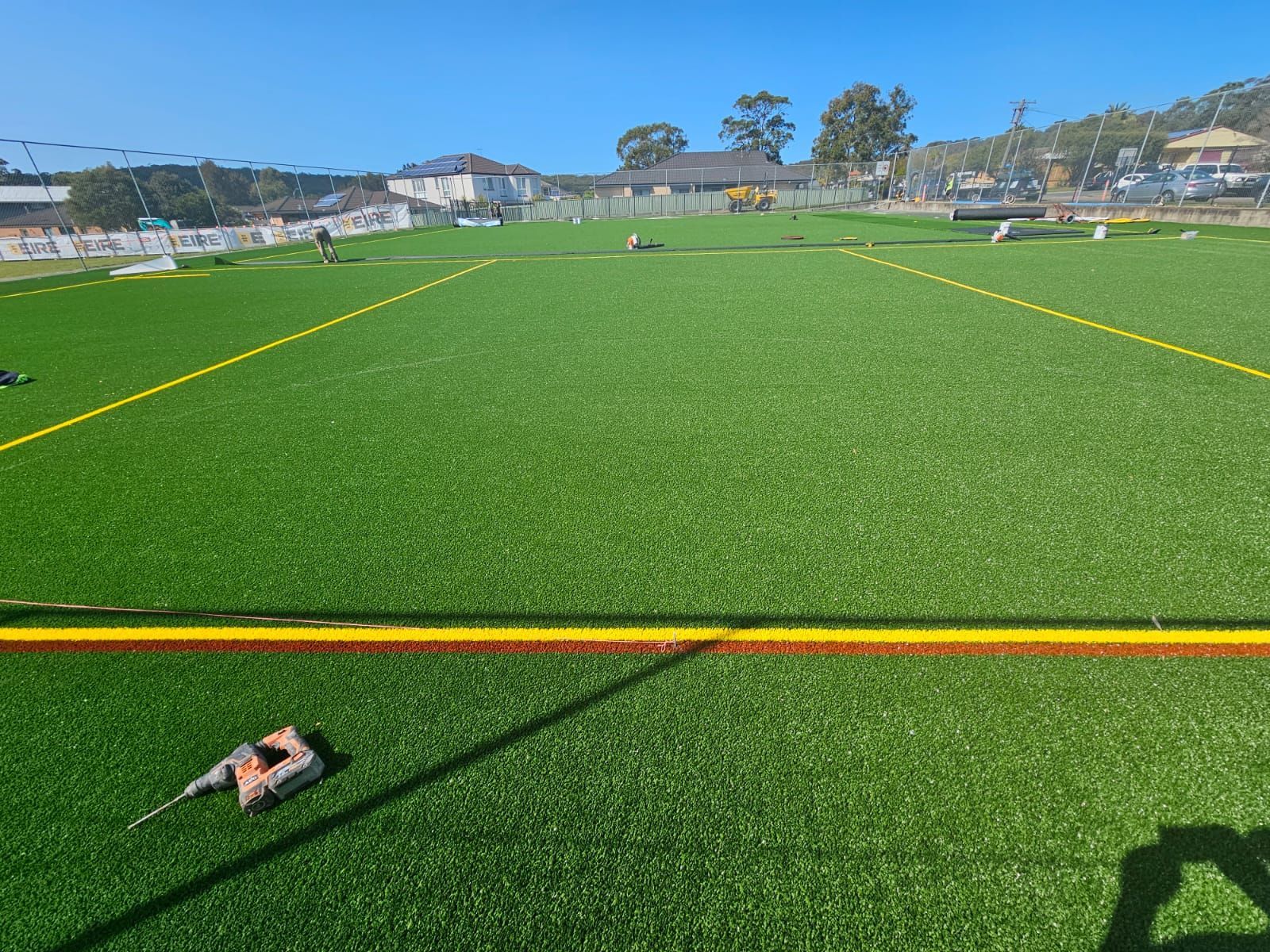 Artificial turf sports field, freshly installed, with yellow lines, in daylight. Construction equipment visible in the background.