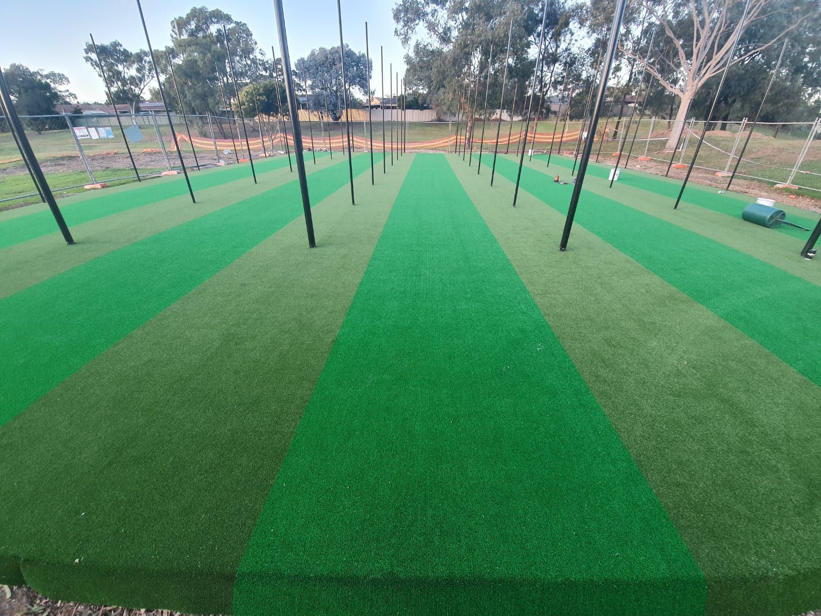 Artificial turf field with alternating light and dark green stripes, black poles. Outdoors with trees in the background.