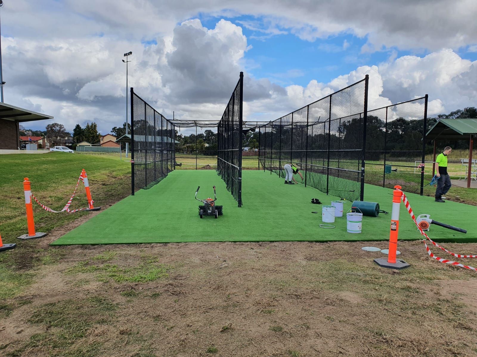 Cricket practice nets with artificial turf, surrounded by black fencing. A worker is present.