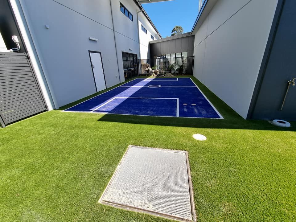 A courtyard with a blue painted sports court surrounded by green turf and two buildings.