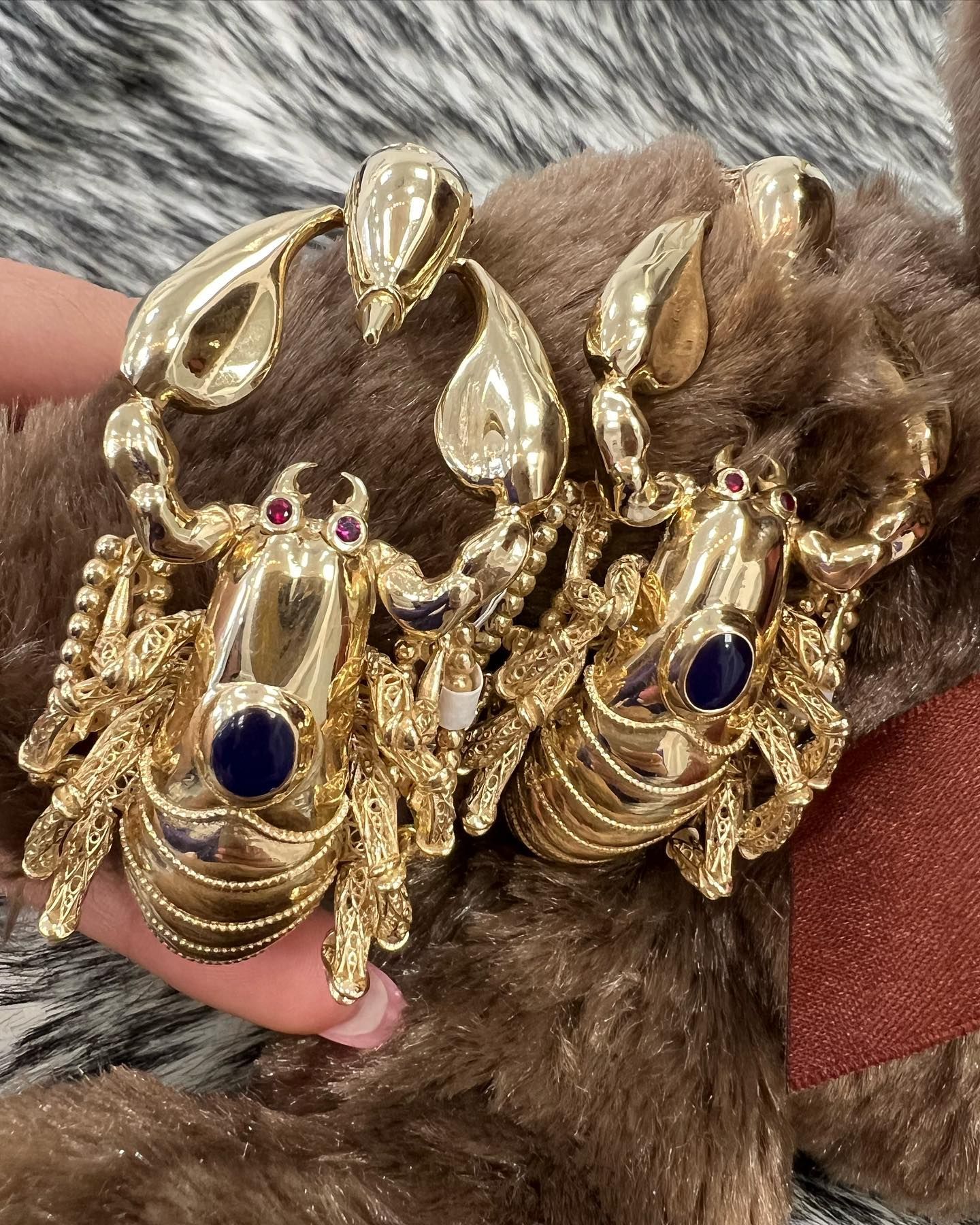 Una persona sostiene un par de pendientes de escorpión de oro. Yene Joyeria