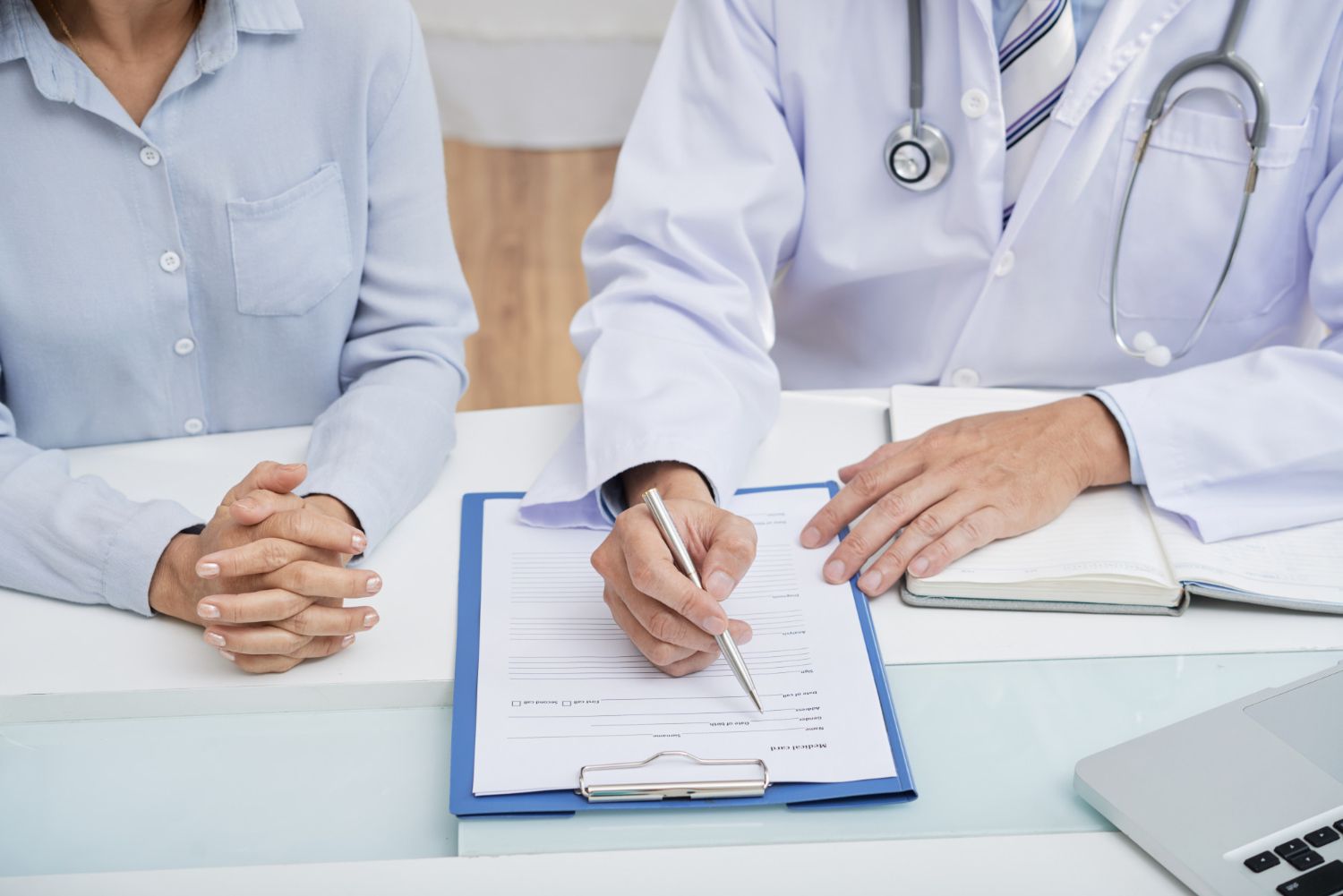 A doctor and a patient are sitting at a table and the doctor is writing on a clipboard.
