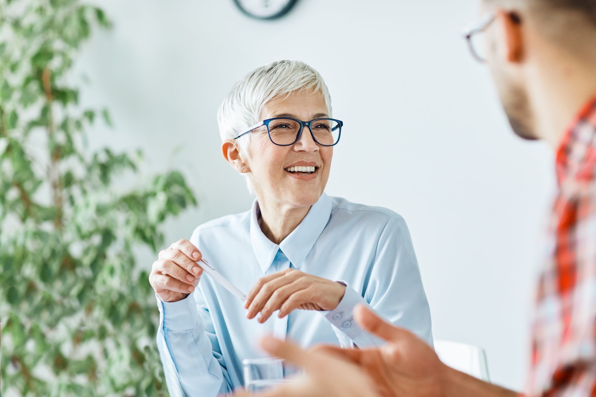 A person with short light hair and glasses smiles while talking to someone out of frame in a bright office space.