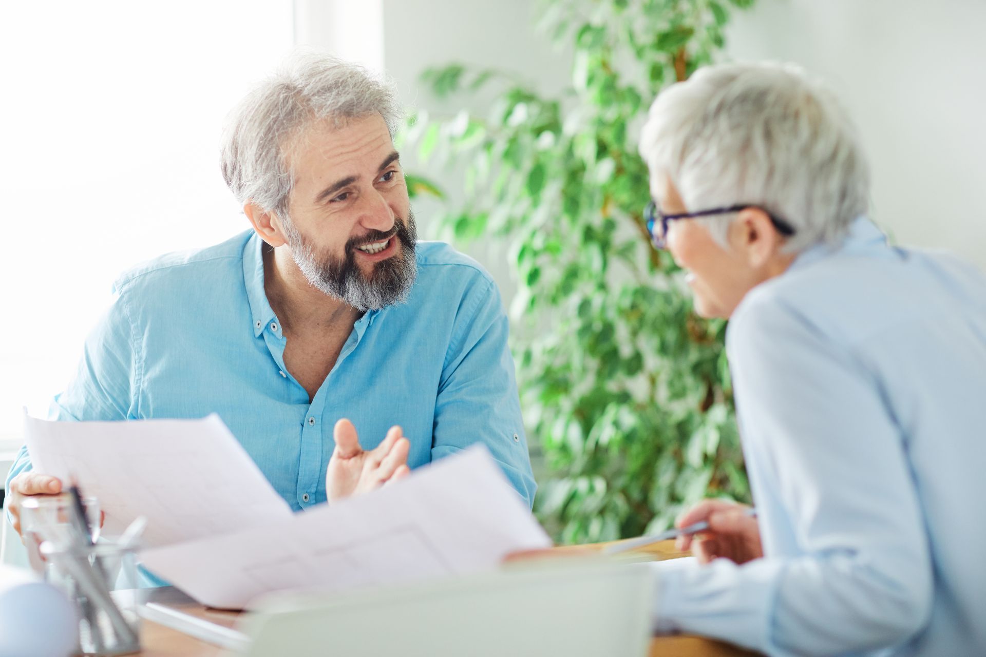 Two people sitting at a table with papers, engaged in a discussion in a brightly lit, plant-filled office.