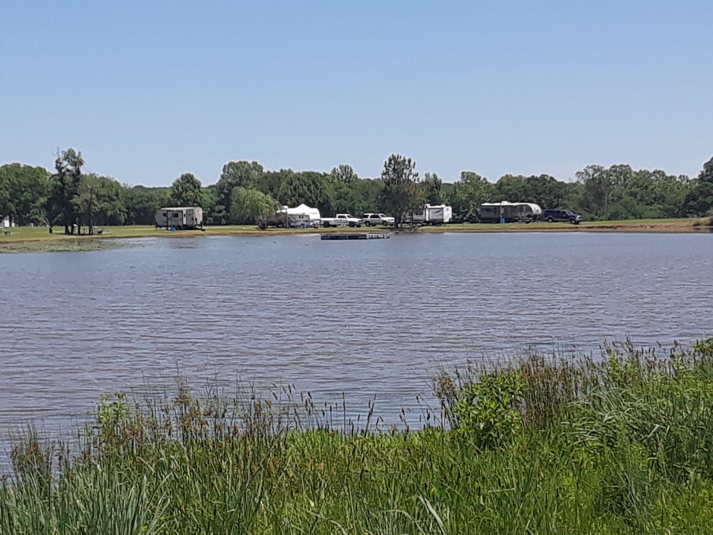 Lake view: campers and trees on the far shore under a blue sky, tall grass in foreground.