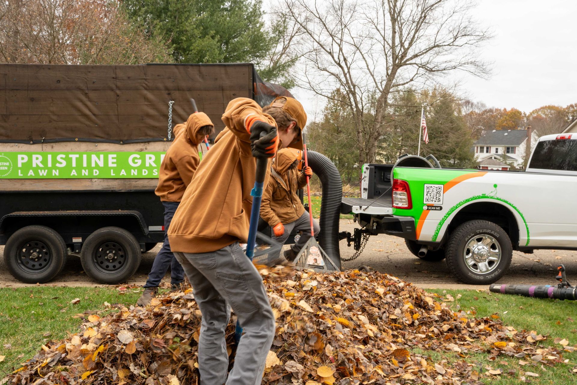Landscapers in brown hoodies loading leaves with shovels and a vacuum into a trailer.