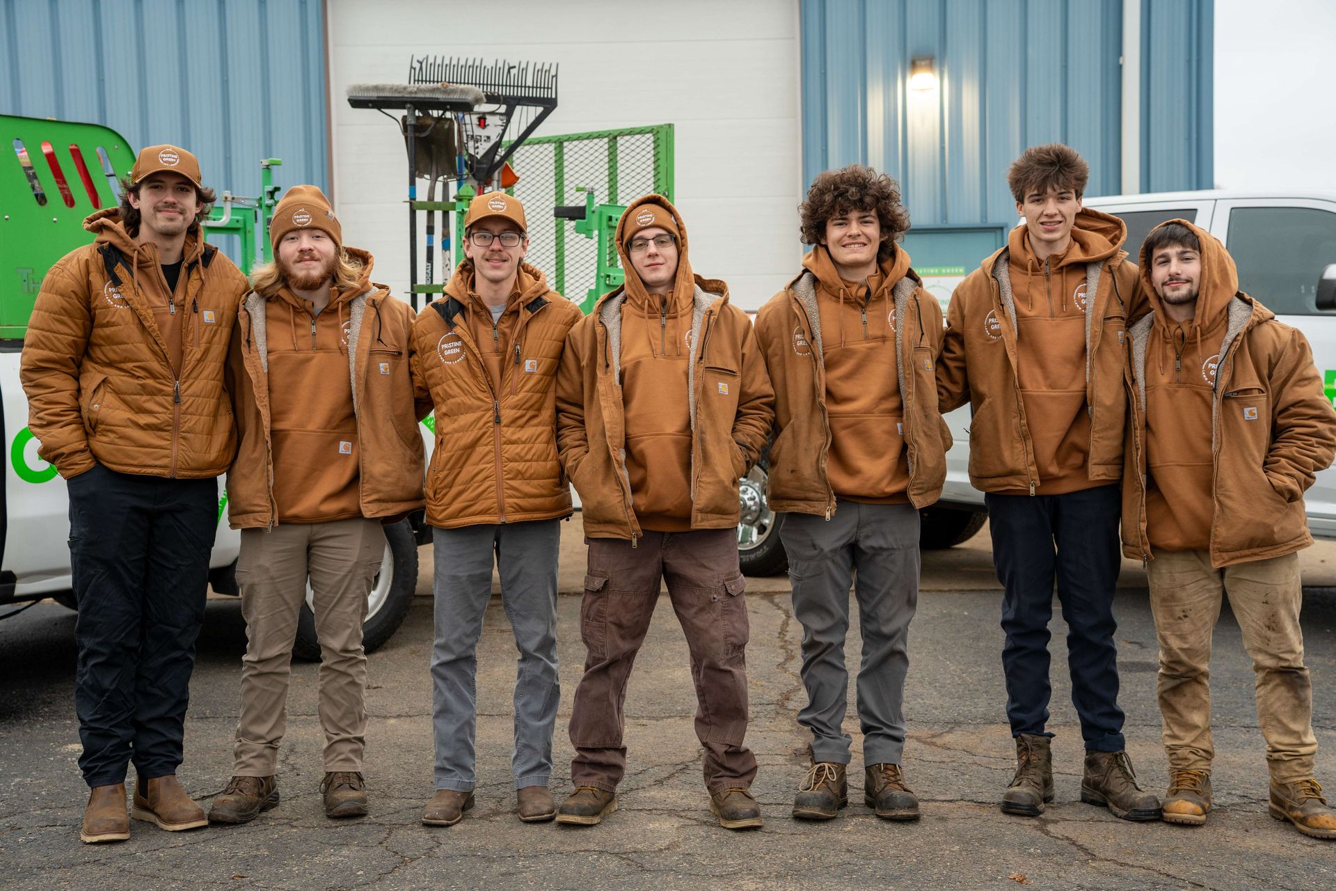 Seven people in brown jackets and hats stand outside, smiling. They are posing by green equipment and a white truck.