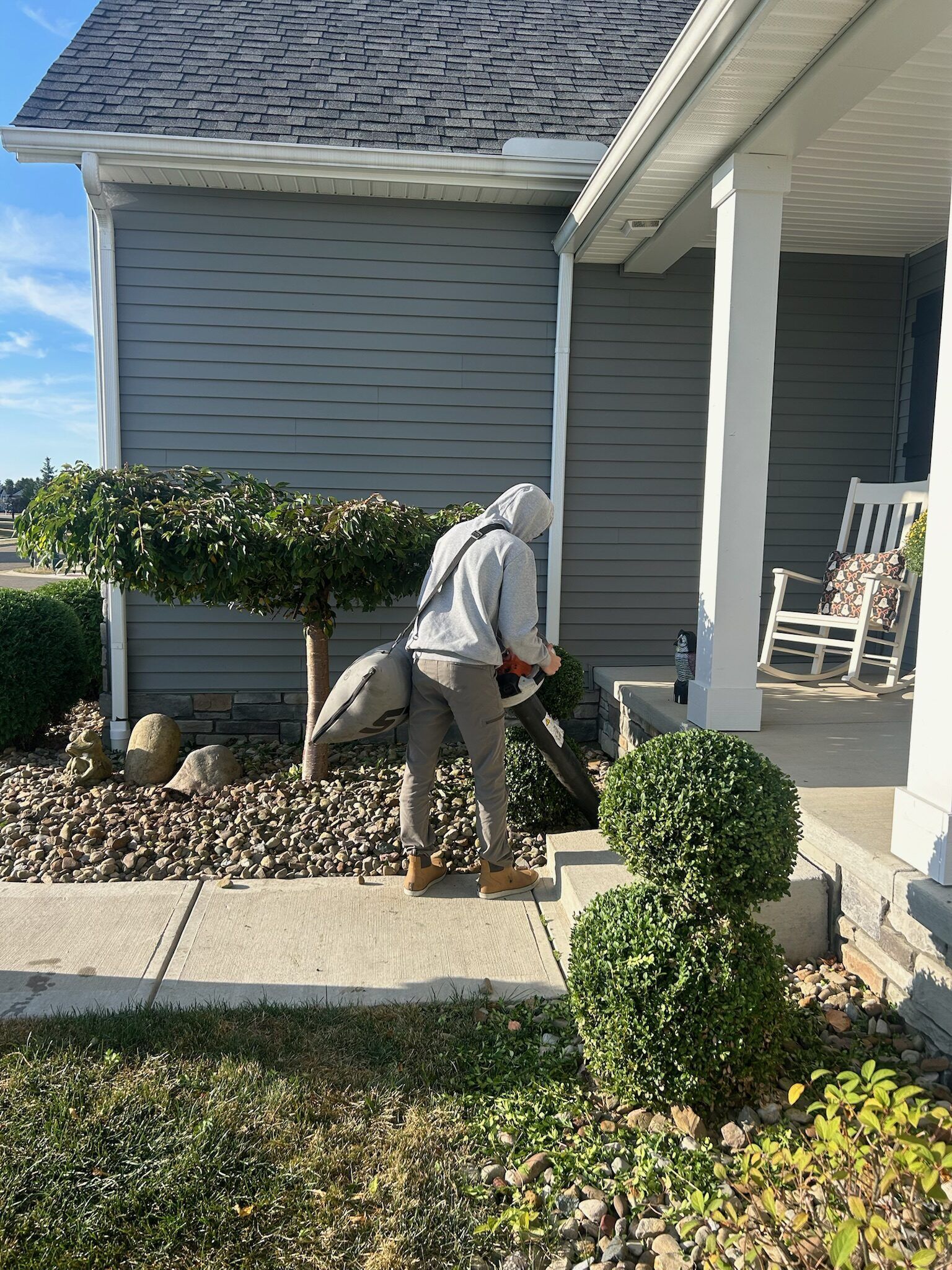 Person blowing leaves next to a house with gray siding and a porch.