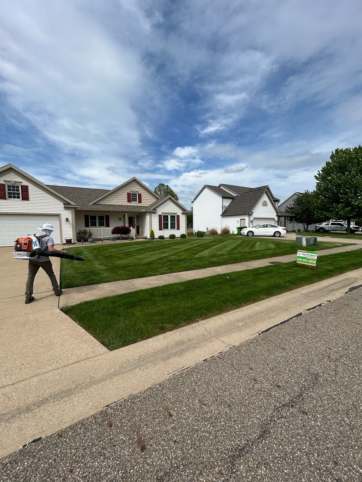 Person using a leaf blower on freshly cut green grass in a suburban neighborhood under a cloudy blue sky.